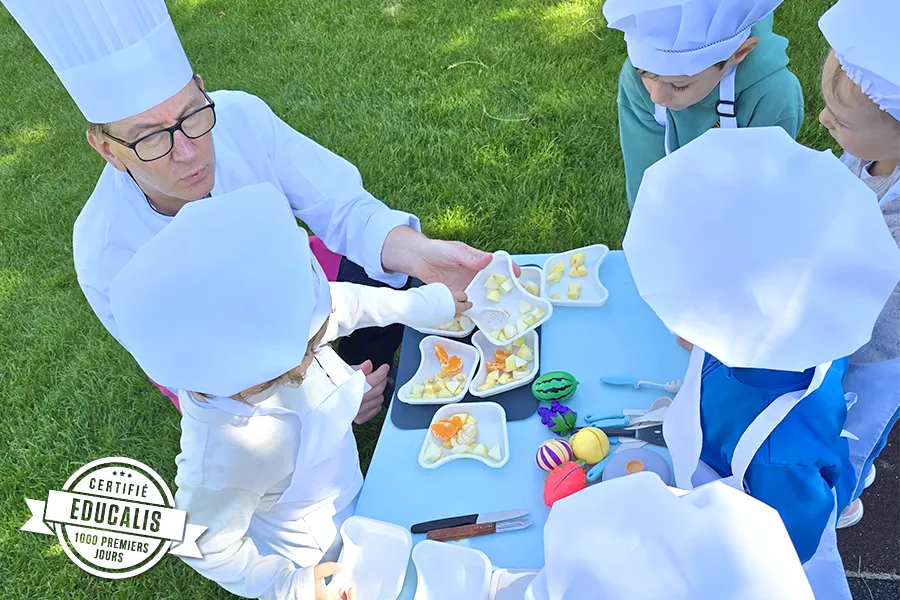 Enfants du groupe des Grands en pleine préparation de leur salade de fruits à la crèche 6 Sens, encadrés par un professionnel de Novae à Saint-Sulpice.